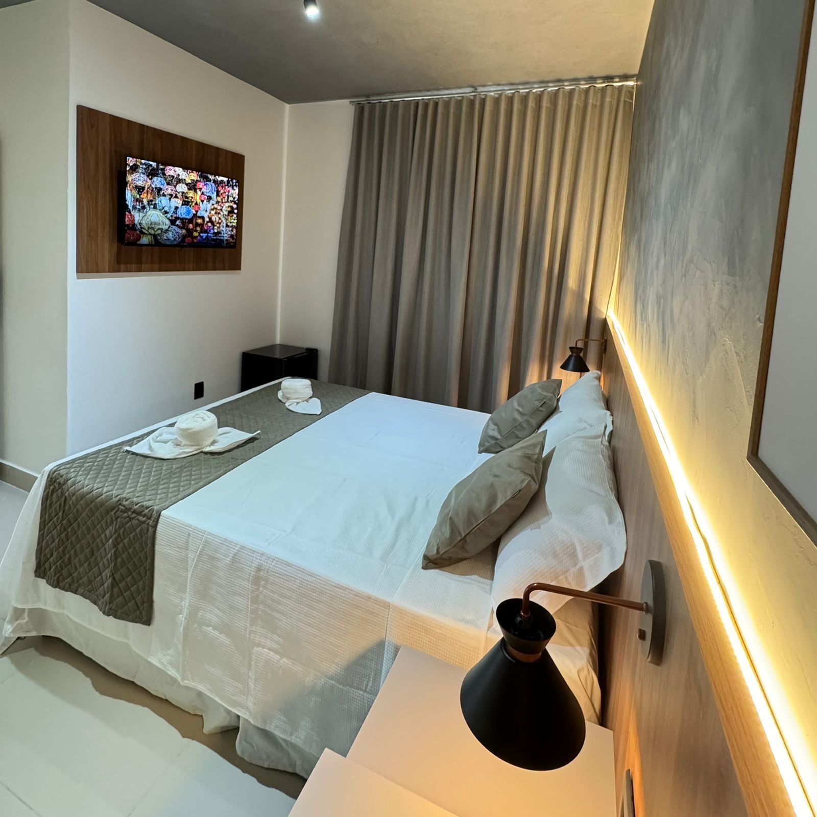 A modern hotel room with a large bed, a flat-screen TV mounted on the wall, a bedside table with a lamp, and a window with curtains.
