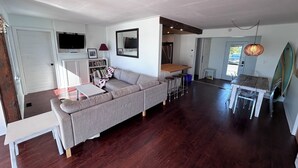 Un salon moderne avec un canapé d’angle gris, une table basse blanche et un coin repas comprenant une table en bois et des tabourets de bar.