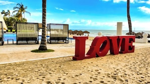 Una spiaggia con grandi lettere rosse che compongono la scritta 'LOVE' e un'area piscina con lettini e ombrelloni.