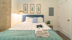 Een net opgemaakt bed met een blauw hoofdeinde, een bedlampje en een klein tafeltje met een plant.