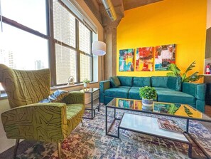 A modern living room with a green sofa, a patterned armchair, and a coffee table. There are large windows with a city view, a yellow accent wall, and a floor lamp.