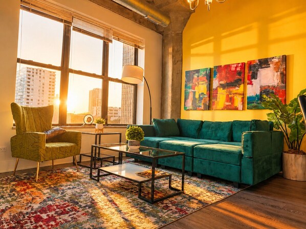 A modern living room with a green sofa, a patterned rug, and large windows with blinds.