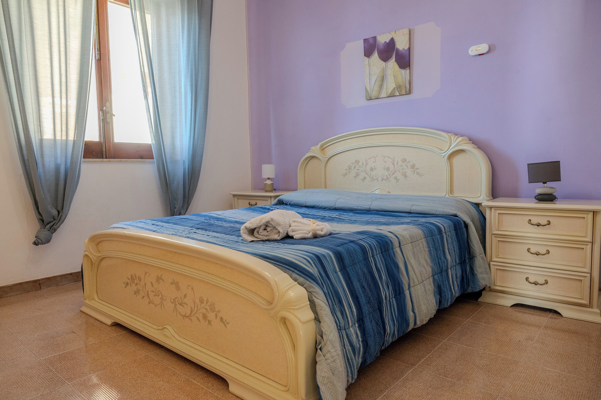 A bedroom with a bed, a nightstand, a dresser, a window with curtains, and a wall-mounted artwork.