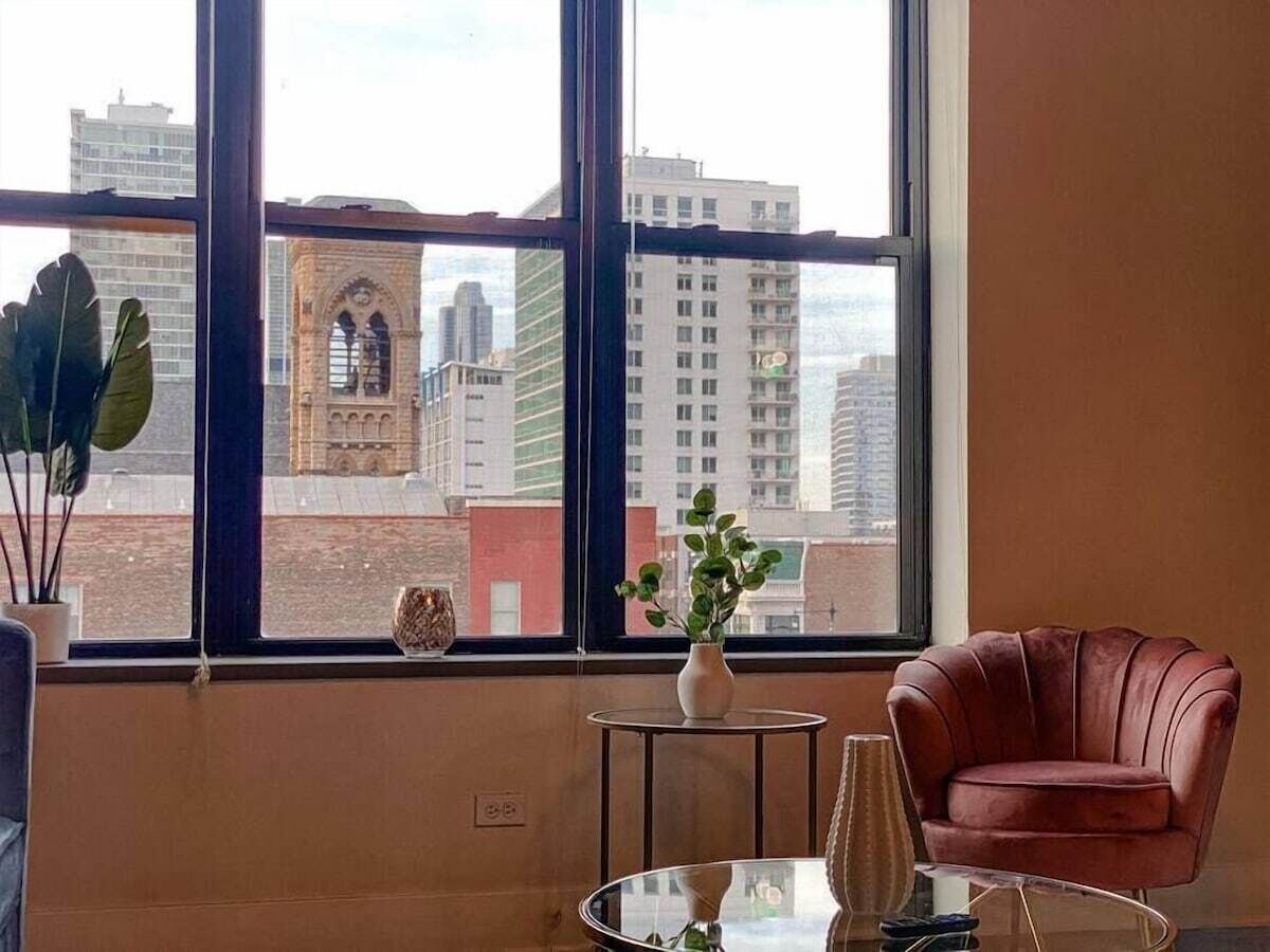 A room with a large window, a red chair, a small table with a vase, and a potted plant.
