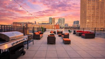 A rooftop patio with outdoor furniture, a grill, and string lights against a sunset cityscape.