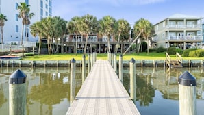 A waterfront promenade with a wooden walkway, palm trees, and buildings in the background. - Laguna Bella B5 | Pool View (Gulf Shores)