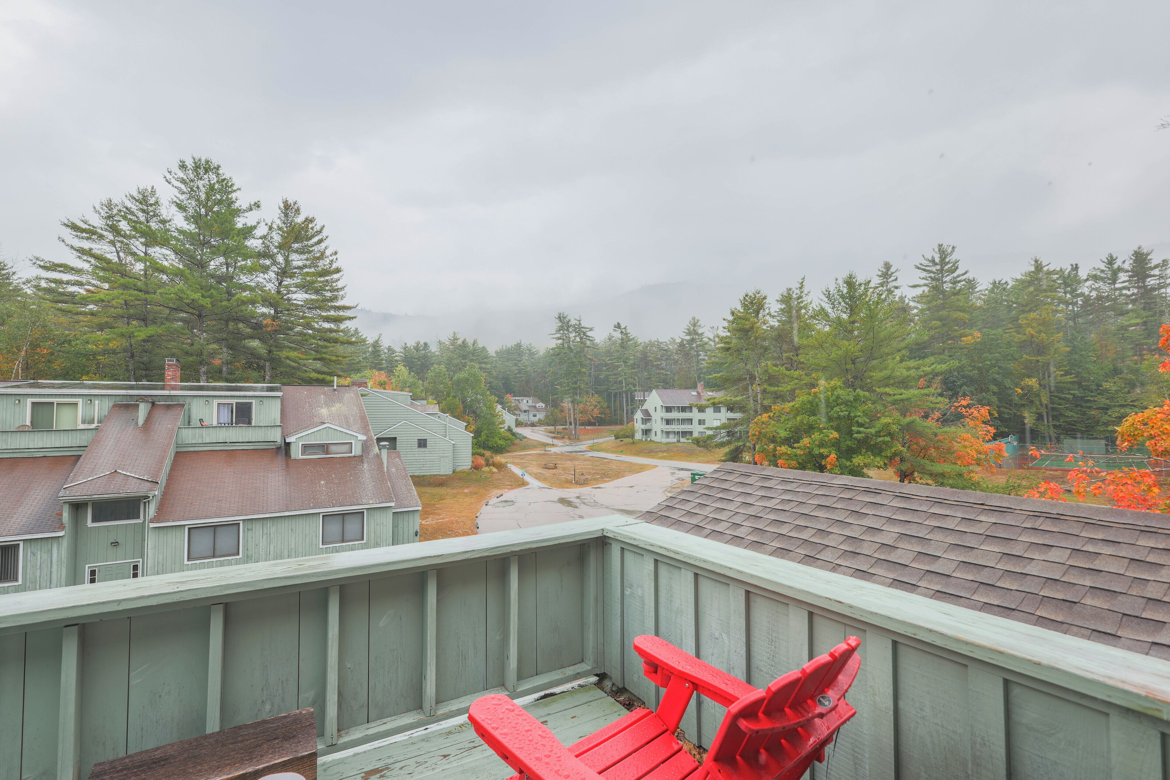 A view from a balcony with a wooden railing, overlooking a residential area with houses and trees.