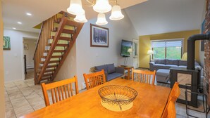 Een eetgedeelte met een houten tafel, stoelen en een decoratieve schaal. Een trap met houten leuningen leidt naar een andere kamer. Een bank en een televisie staan op de achtergrond.