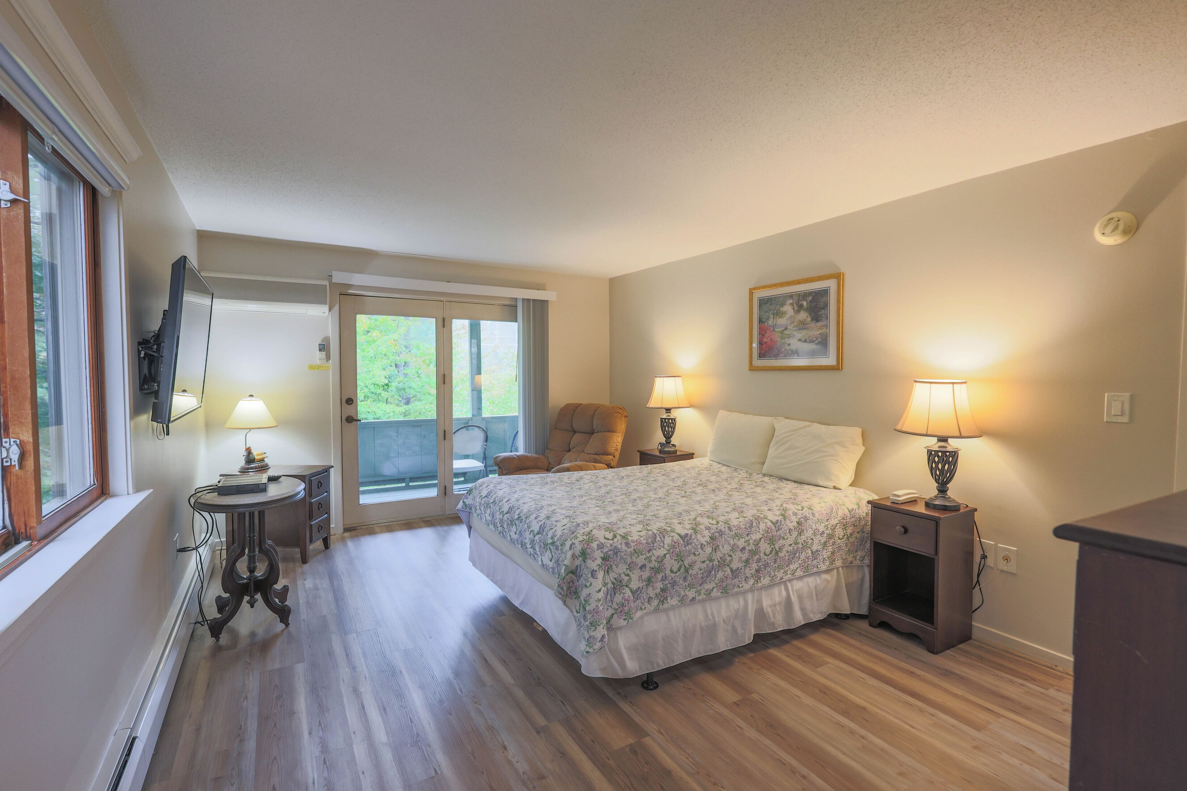 A bedroom with a bed, two bedside tables with lamps, a chair, a window, and a door leading to a balcony.