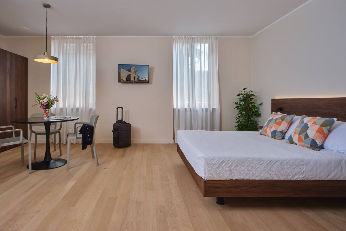 A hotel room with a wooden floor, a bed with patterned pillows, a desk with a chair, a table with a vase of flowers, a suitcase, and a painting on the wall.
