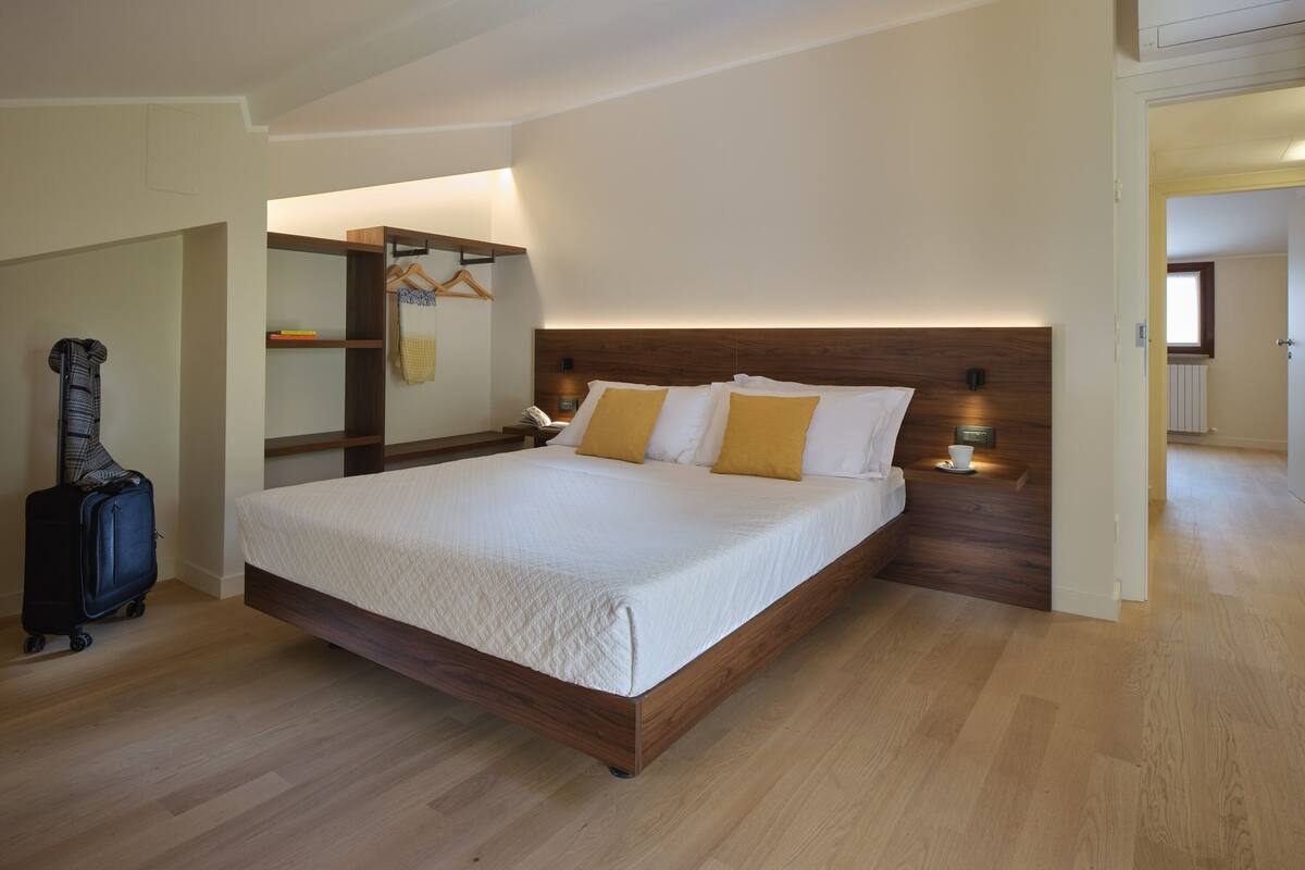 A hotel room with a large bed, wooden headboard, bedside lamps, a suitcase, and a coat rack.