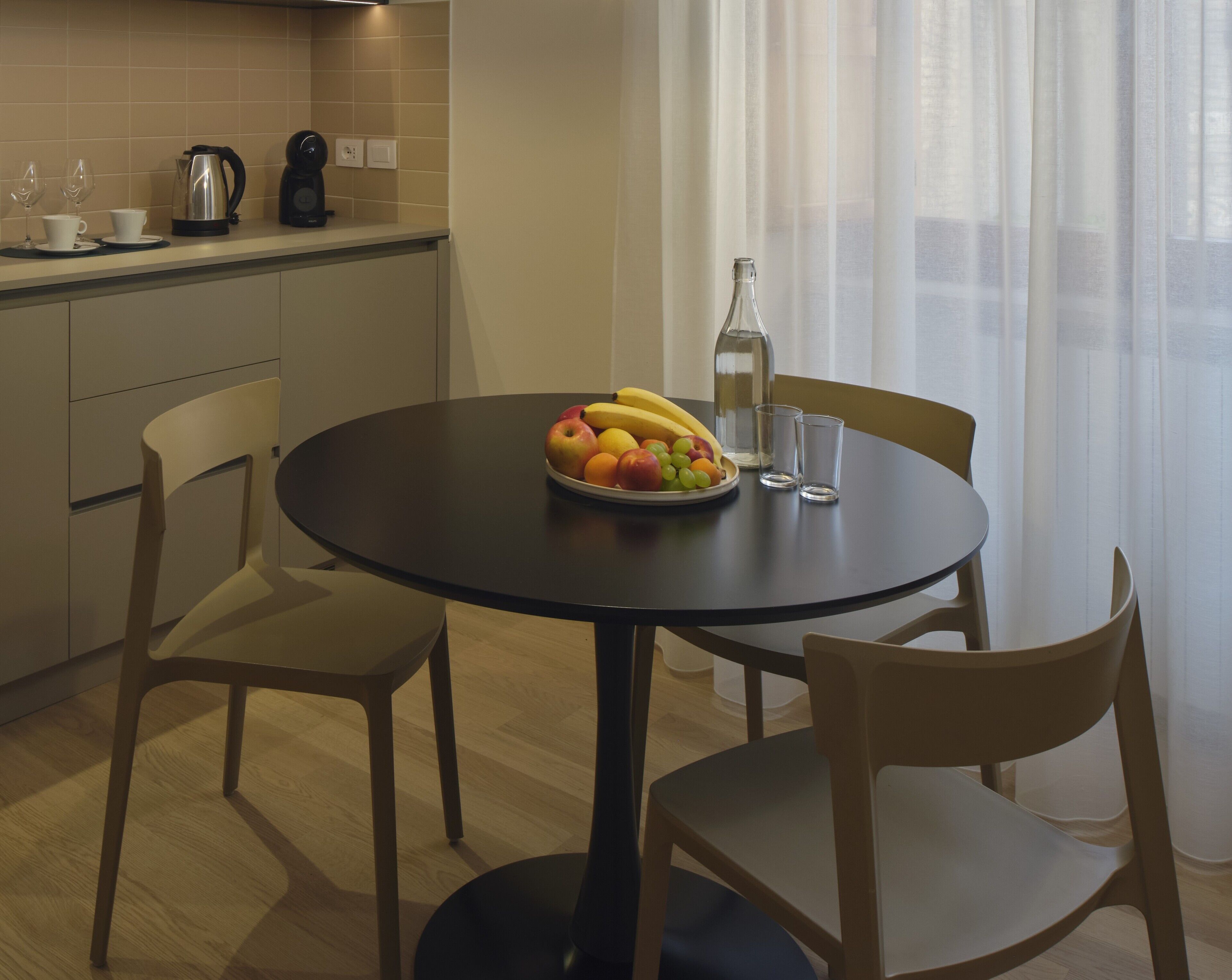 Un tavolo rotondo da pranzo con una ciotola di frutta, due tazze, una macchina da caffè e una bottiglia.