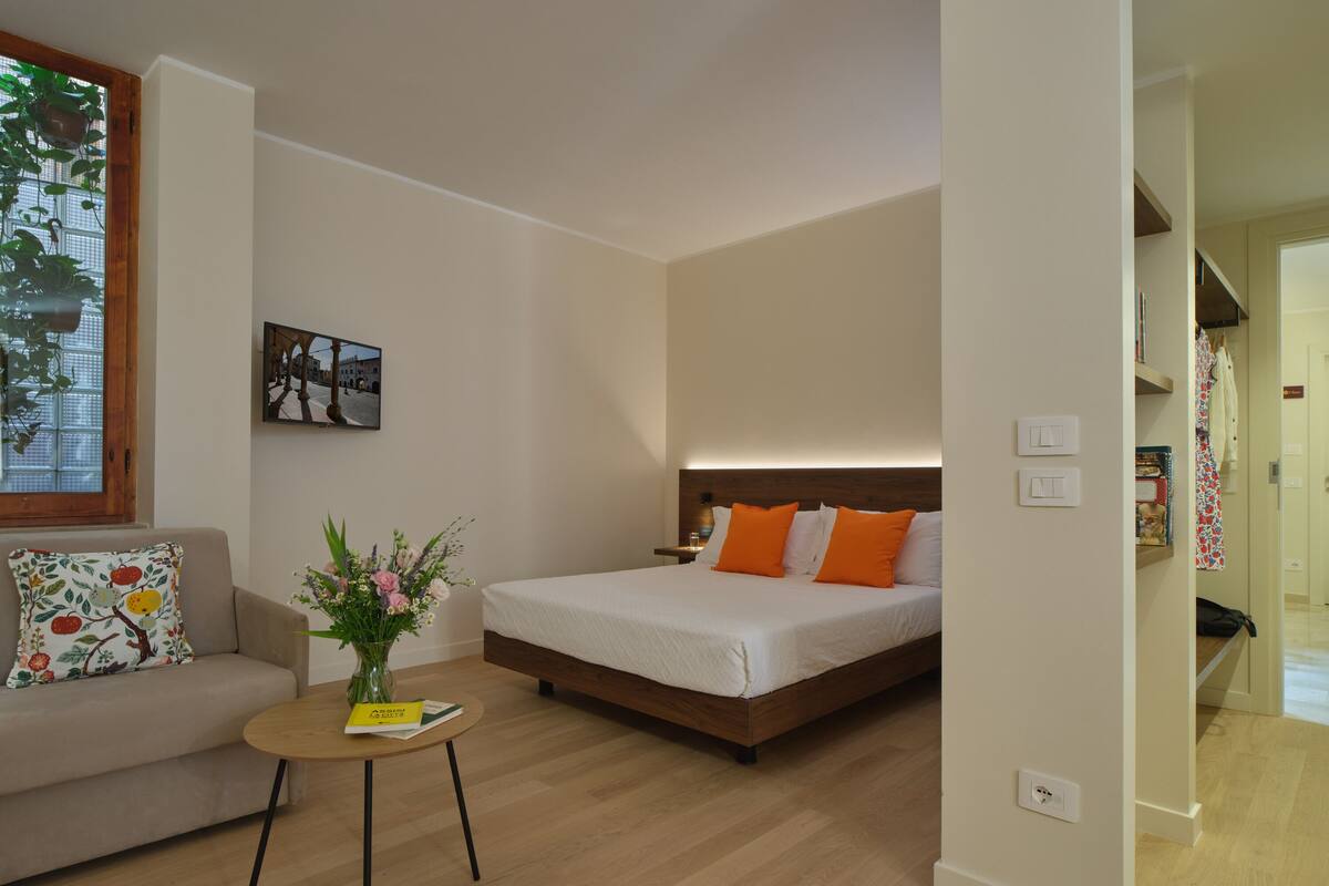 A modern bedroom with a wooden bed, a small round table with a vase of flowers, a sofa with a floral cushion, and a picture on the wall.