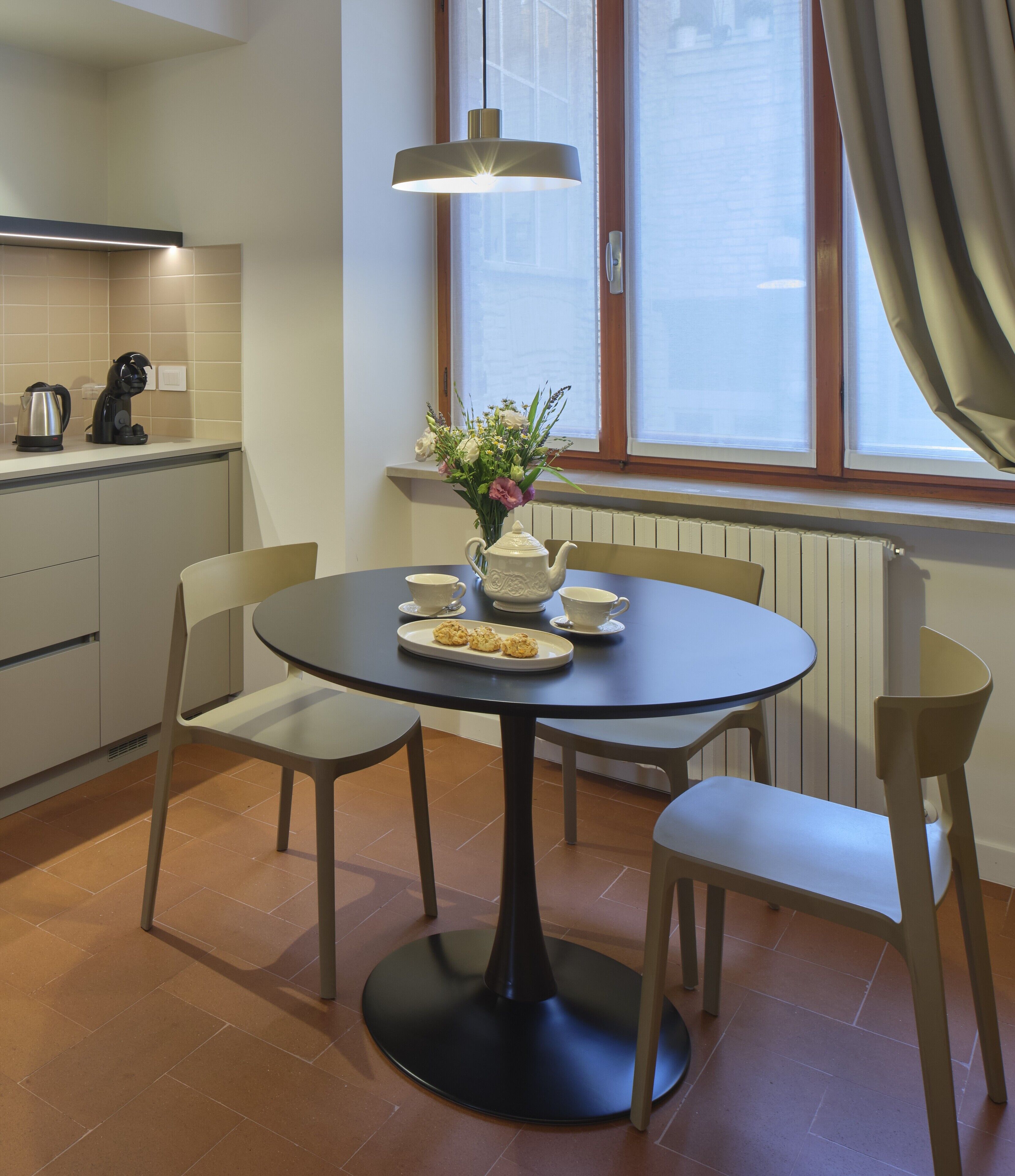 Una cocina moderna con una mesa redonda de comedor, sillas, un hervidor, una tetera, tazas y una bandeja con galletas.