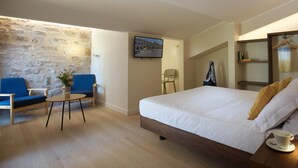 Une chambre d’hôtel avec un mur en pierre, un lit avec une literie blanche, un fauteuil bleu, une petite table sur laquelle est posé un vase de fleurs et un téléviseur fixé au mur.