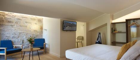 Habitación de hotel con pared de piedra, cama con ropa de cama blanca, sillón azul, pequeña mesa con un jarrón de flores y un televisor en la pared.