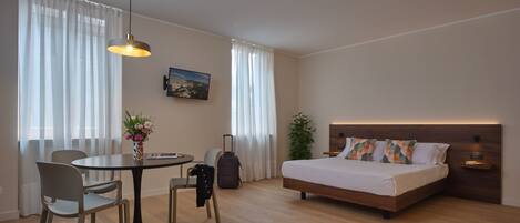 Una habitación de hotel moderna con piso de madera, una cama con cabecera de madera, una mesa de comedor con sillas, una mesita con un jarrón de flores y un ventanal con cortinas translúcidas.