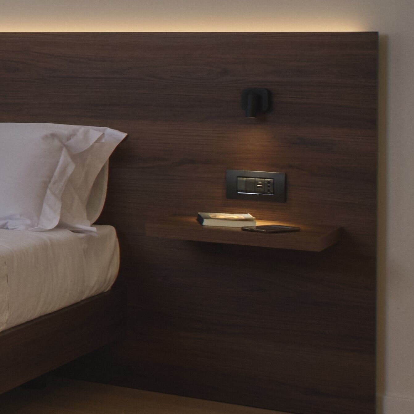 A bedroom with a wooden headboard, a bedside table with a book, and a wall-mounted light fixture.