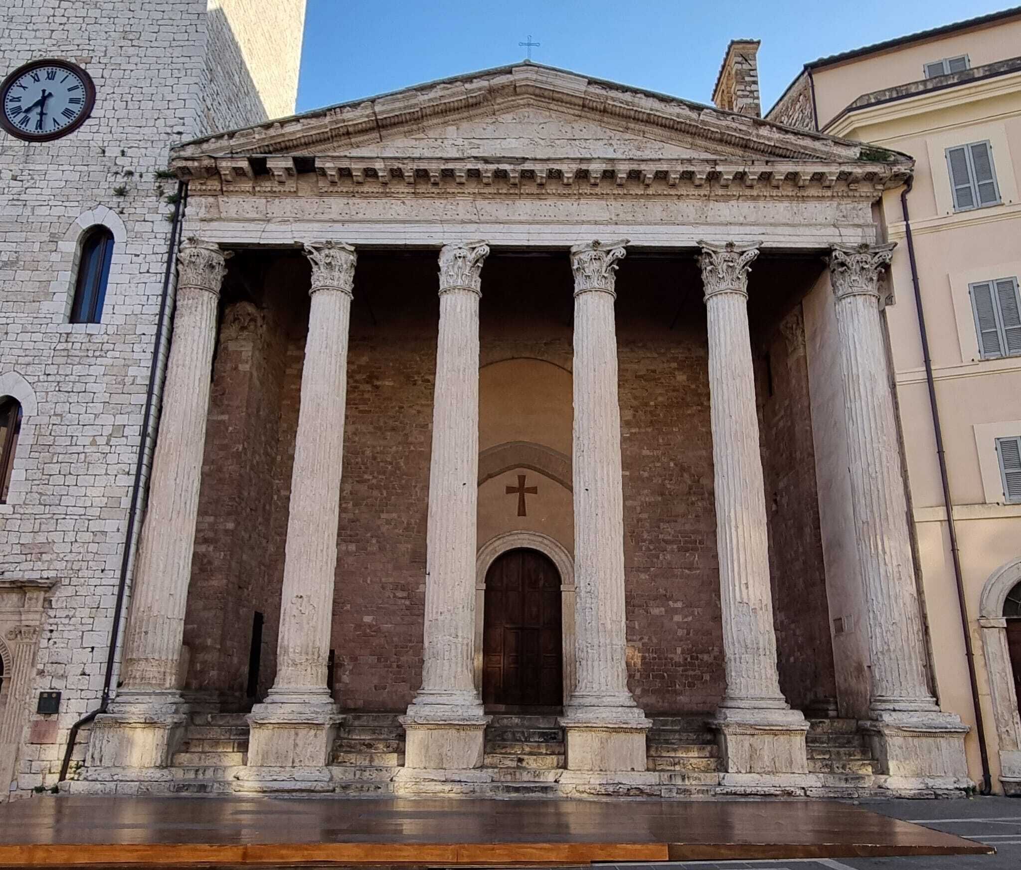 A classical building with a central doorway flanked by columns, a clock on the left, and a cross above the entrance.