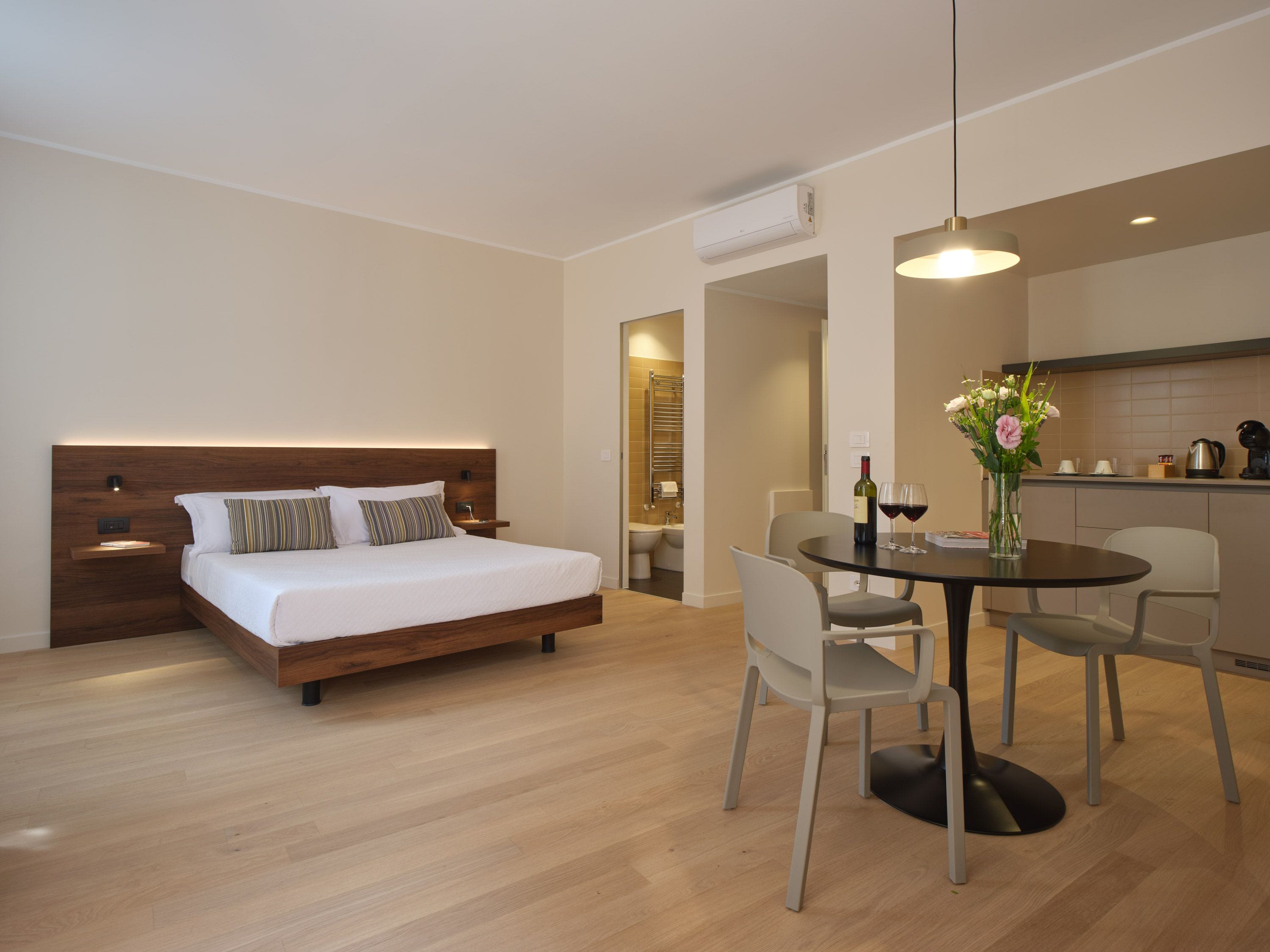 A modern hotel room with a wooden bed, a dining table with chairs, and a kitchen area.