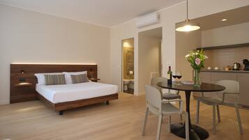 A modern hotel room with a wooden bed, a dining table with chairs, and a kitchen area.
