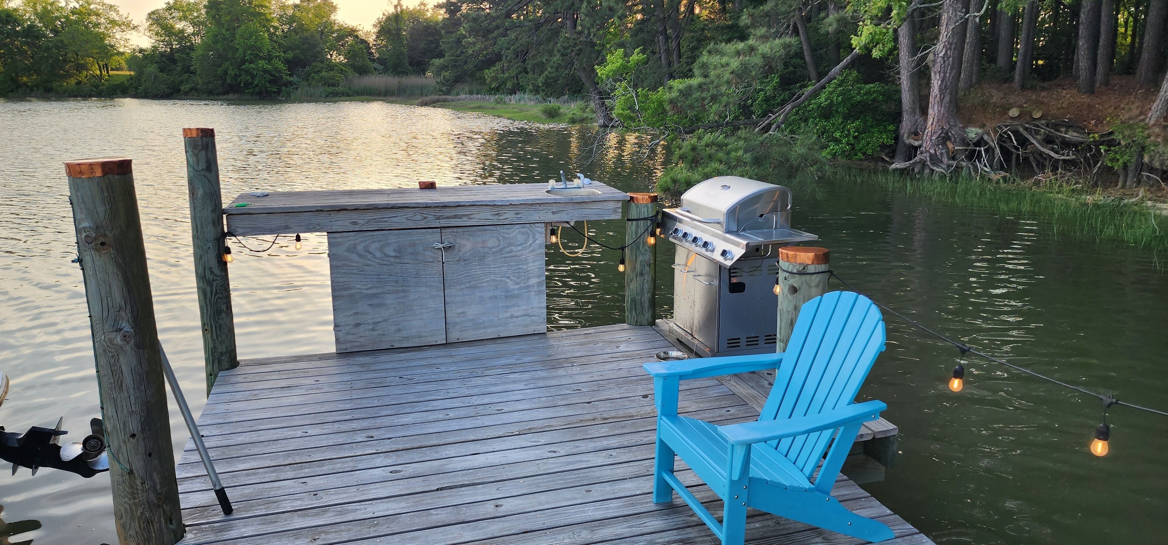 A wooden dock with a grill and a blue chair, overlooking a calm lake.