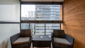 A balcony with wicker chairs and a table, large windows with a city view, and a wooden wall.