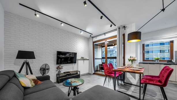 A modern living room with a grey sofa, a dining table with red chairs, a TV mounted on a white brick wall, and a large window offering a city view.