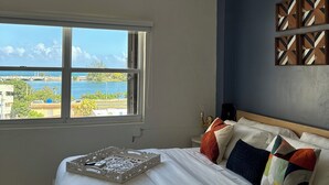 A hotel room with a bed, a ceiling fan, an air conditioning unit, a window with a view of a body of water and buildings, and a wall with a geometric patterned artwork. - Renovated & Authentic San Juan 1-Bedroom APT w/ Caribbean Views (San Juan)