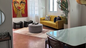 A modern living room with a yellow sofa, a round ottoman, and a glass dining table. There is a large painting on the wall and a potted plant by the window. - Renovated & Authentic San Juan 1-Bedroom APT w/ Caribbean Views (San Juan)