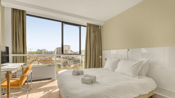 Ein Hotelzimmer mit Bett, Schreibtisch und Stuhl. Ein großes Fenster mit Vorhängen bietet Blick auf die Stadt.
