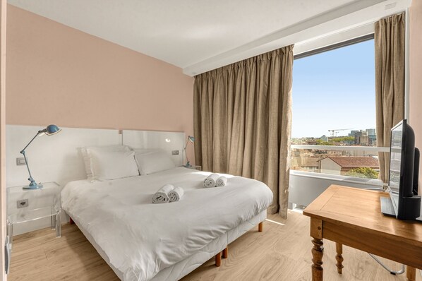 A hotel room with a large bed, a wooden desk, a television, and a window with curtains.