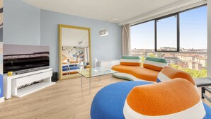 Ein modernes Wohnzimmer mit großem Fenster, Flachbildfernseher, Glastisch und farbenfroher Sitzgruppe.