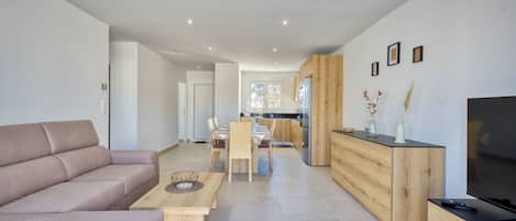 A modern living room with a sofa, a dining table, and a kitchen area with wooden cabinets and a TV.