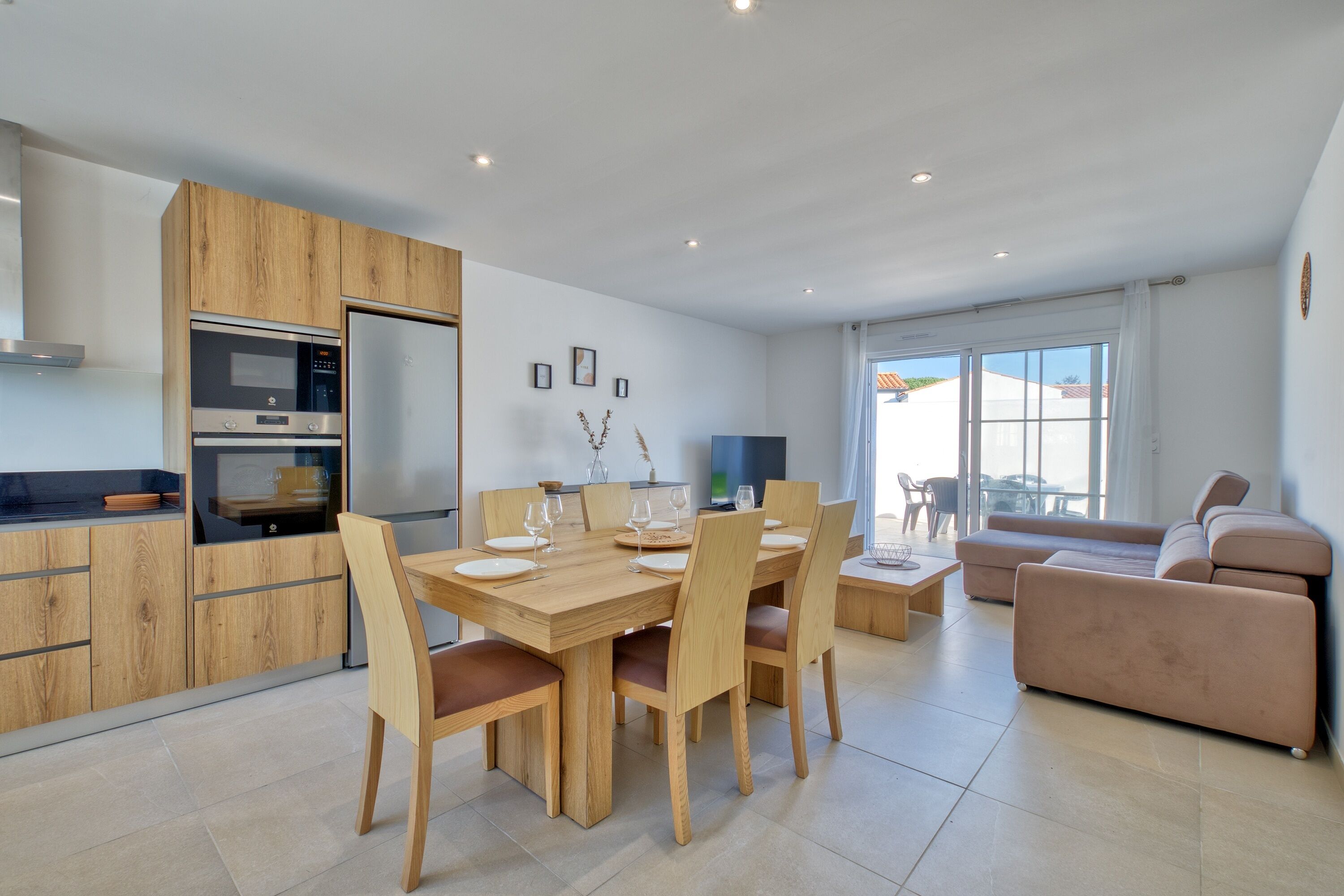 A modern kitchen with wooden cabinets and a built-in oven. There is a dining area with a wooden table and chairs. A sofa and a TV are visible in the living room.