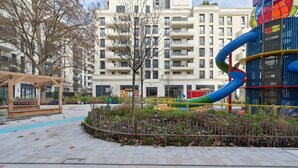 Ein Spielplatz mit Rutsche und Klettergerüsten, umgeben von einer gepflasterten Fläche und einem Gebäude im Hintergrund.