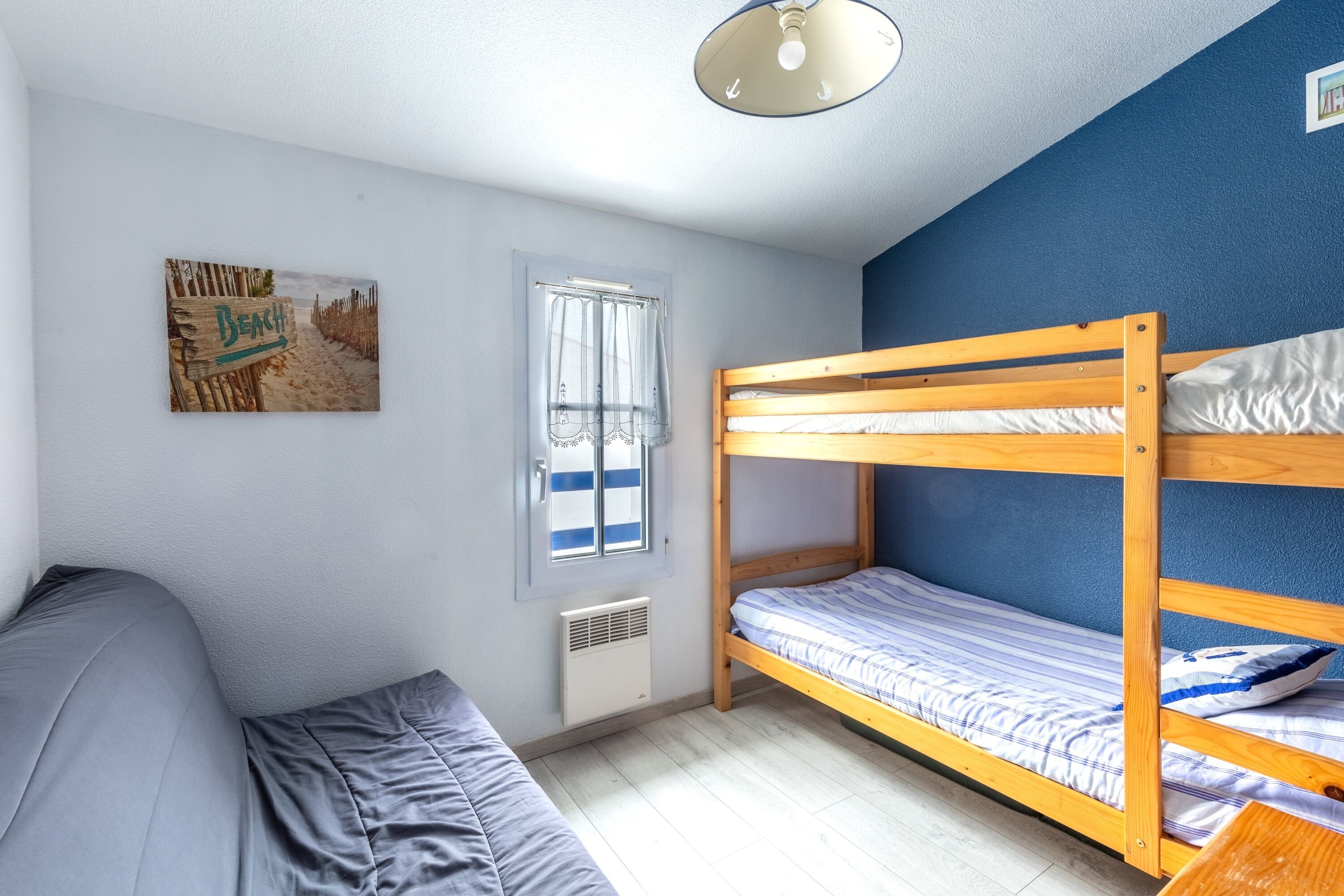 A room with a bunk bed, a sofa, a window with curtains, a wall-mounted picture, and a ceiling light fixture.