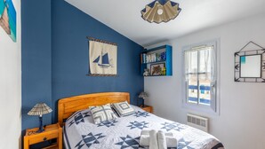 Un dormitorio con cabecera de madera, una cama con colcha de diseño geométrico, dos lámparas de noche, un adorno de velero montado en la pared, una estantería y una ventana con vistas a edificios.