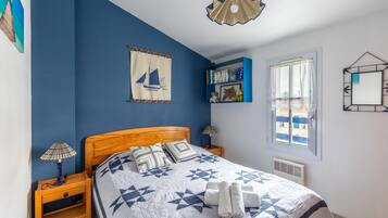 Une chambre à coucher avec une tête de lit en bois, un lit recouvert d’une courtepointe à motifs géométriques, deux lampes de chevet, une décoration murale représentant un voilier, une bibliothèque et une fenêtre donnant sur des immeubles.