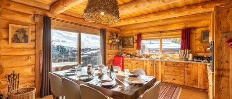 A wooden cabin interior with a dining table set for four, a kitchen area, and a large window offering a view of the outdoors.