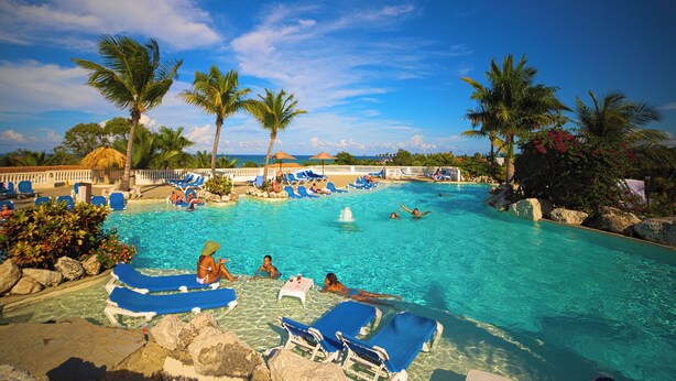 Une grande piscine extérieure à l’eau d’un bleu limpide, entourée de palmiers et de chaises longues.