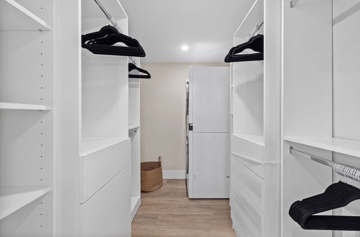 A narrow walk-through closet with white shelving units, a basket, and a refrigerator.