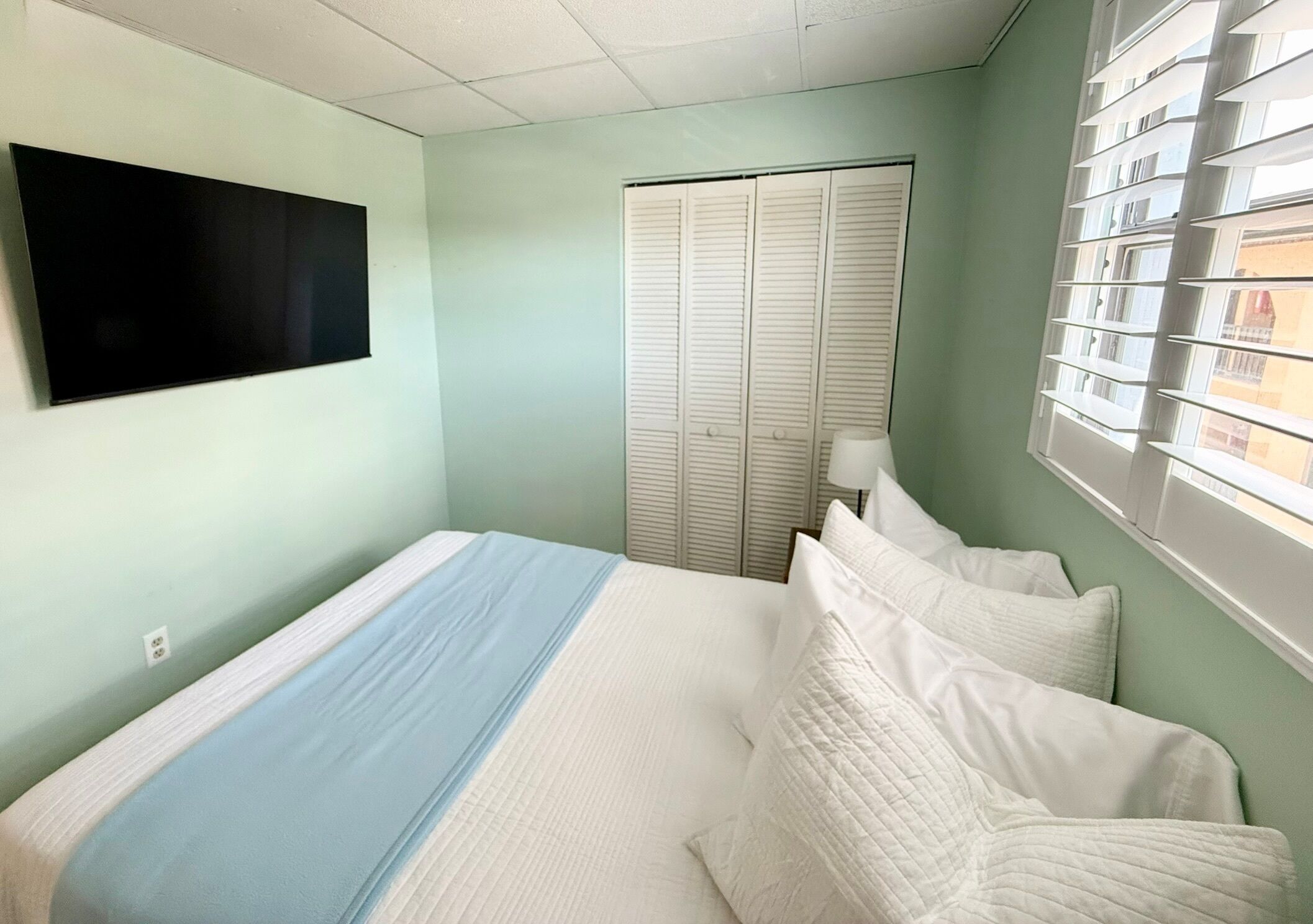 A bedroom with a bed, a television mounted on the wall, a window with blinds, and a closet with sliding doors.