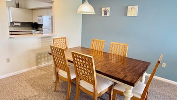 A dining area with a wooden table and chairs, a hanging light fixture, and a kitchen in the background.