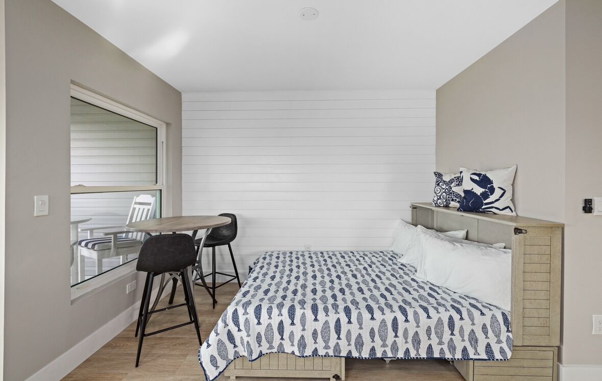 A small, compact bedroom with a single bed, a small table with two chairs, and a window with blinds.