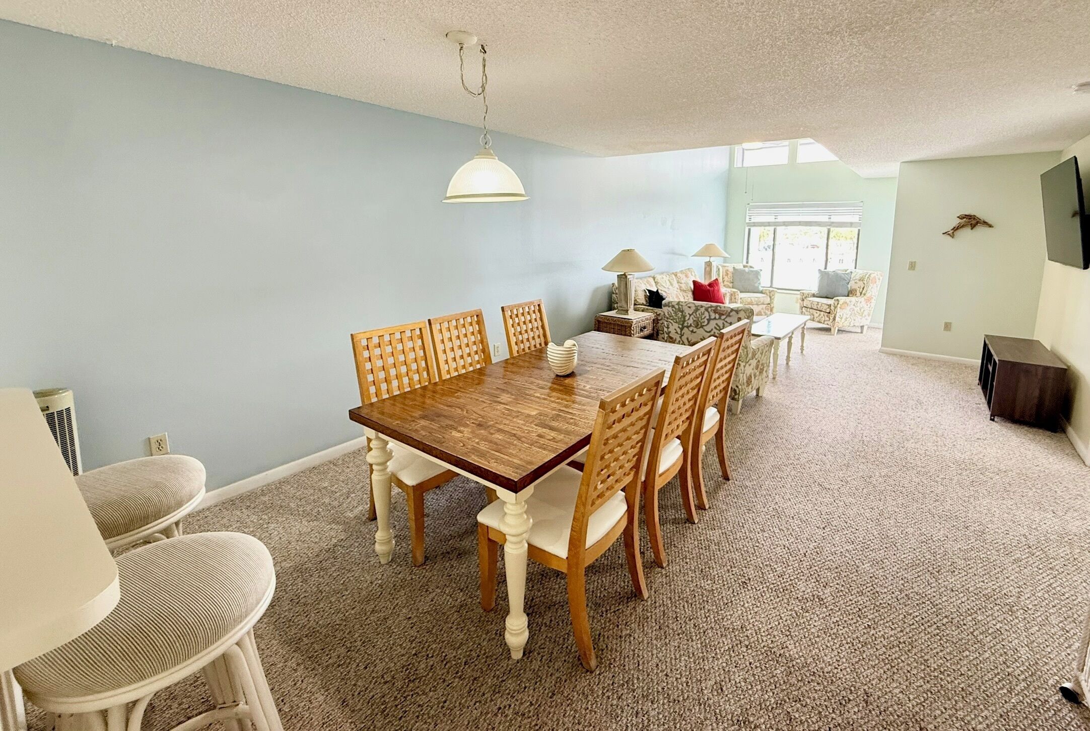 Un comedor con mesa y sillas de madera, una lámpara colgante y vista al exterior a través de una ventana.