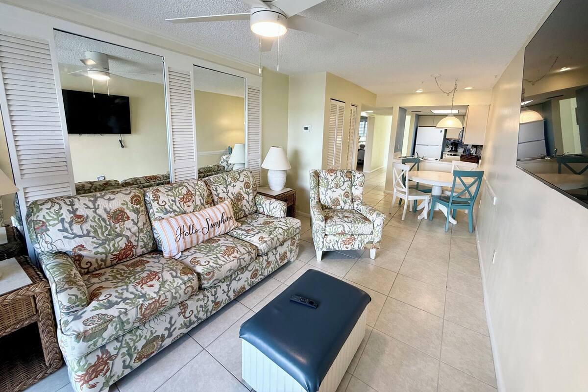 Una sala de estar con sofá, sillón, reposapiés, ventilador de techo, televisor y una zona de comedor con sillas y mesa.