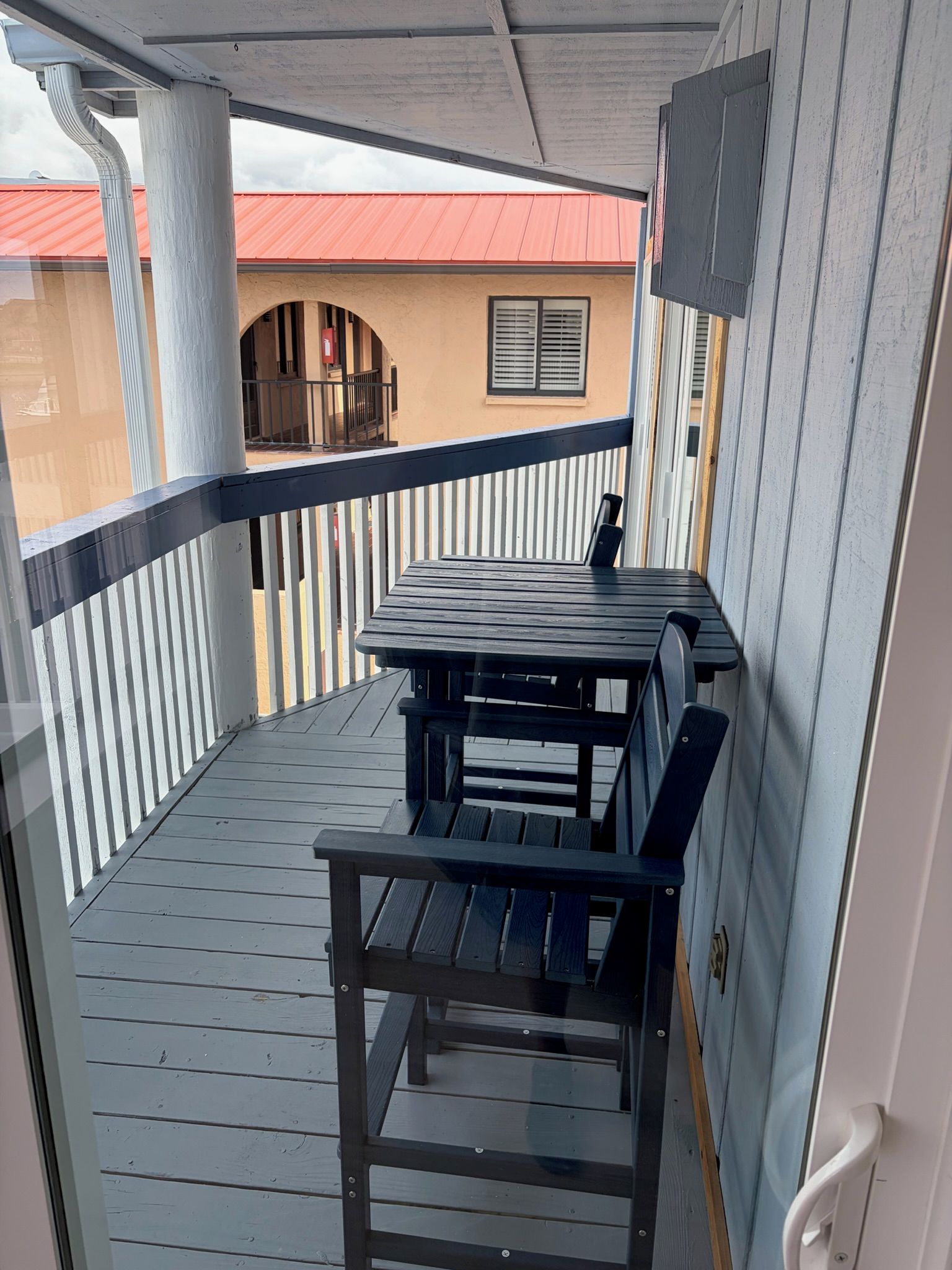 A balcony with a wooden table and chairs, a white railing, and a view of a building with a red roof.
