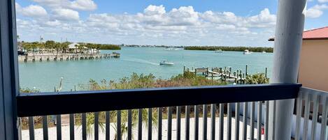 A view from a balcony with a railing, overlooking a calm body of water with boats, a dock, and a distant shoreline with buildings.
