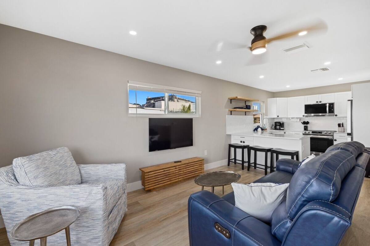 Una sala de estar moderna con un sofá, una mesa de centro y un televisor. Hay una zona de cocina con microondas y refrigerador.